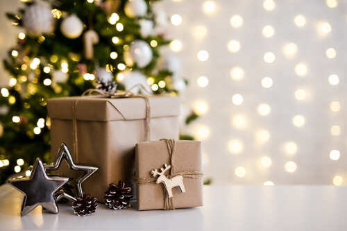 Pacchi regalo decorati con spago e piccoli ornamenti davanti a un albero di Natale illuminato