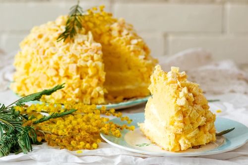 Fetta di torta mimosa con copertura soffice a cubetti su piatto chiaro, decorata con rametti di mimosa fresca