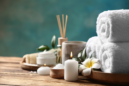 Set benessere con asciugamani arrotolati, candela accesa, sali da bagno e fiore bianco su tavolo in legno.