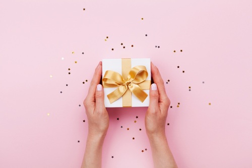 Mani femminili che tengono una scatola regalo con fiocco dorato su sfondo rosa con coriandoli.